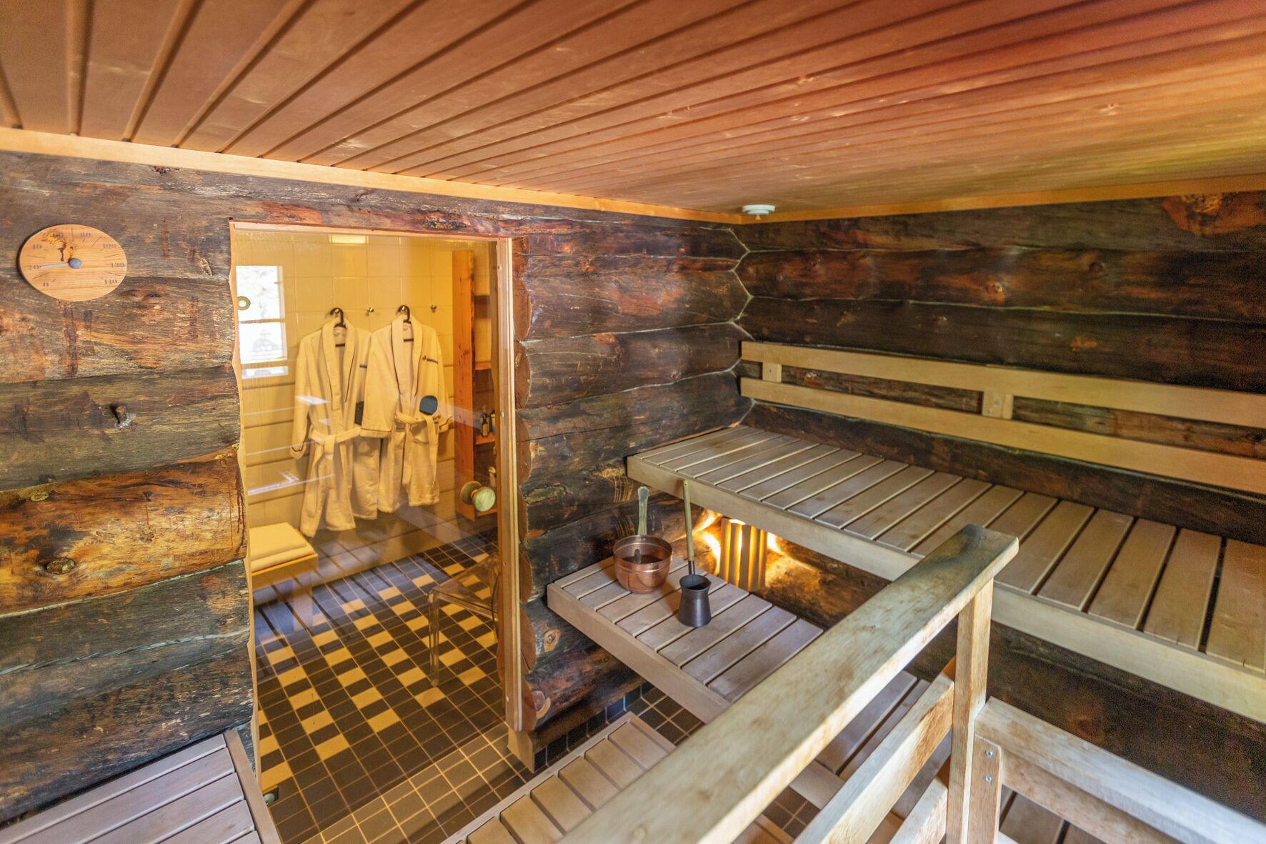 A sauna room with wooden walls, a checkered floor, and a bench. There are towels and a bucket on the bench.