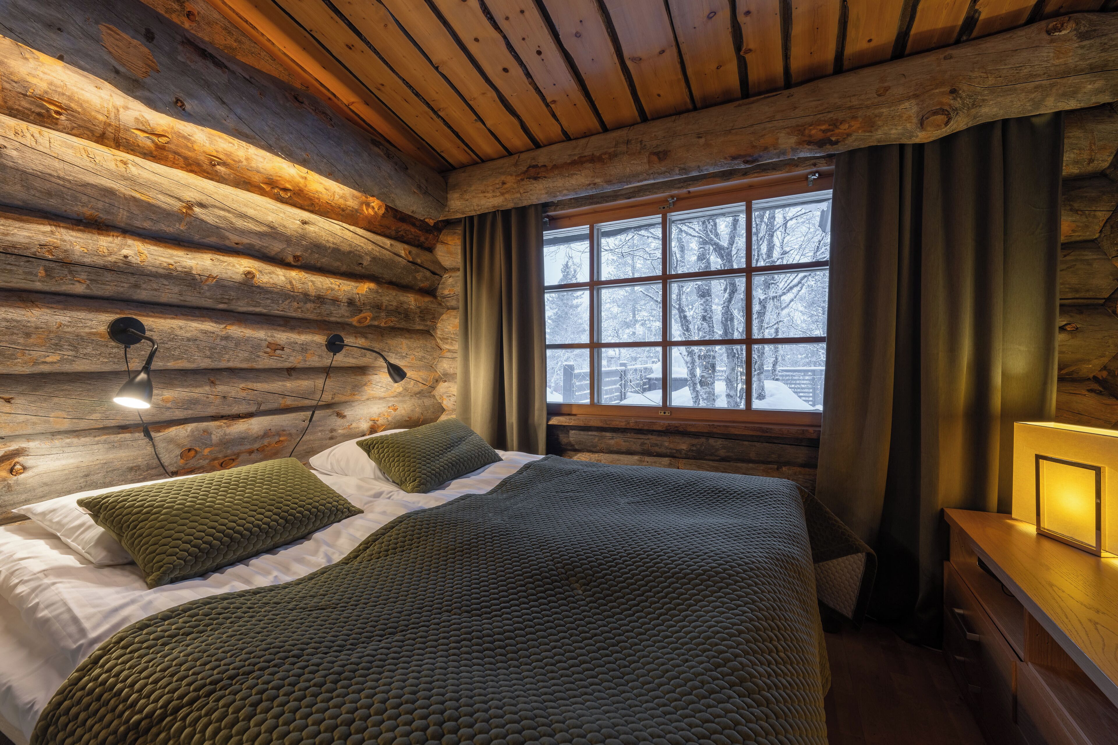 Ein Blockhauszimmer mit einem Bett, zwei Wandleuchten, einem Fenster mit Vorhängen und einem Nachttisch aus Holz mit einer Lampe.