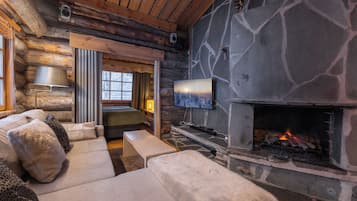 Interior de una cabaña de troncos con chimenea de piedra, un sofá y un televisor.