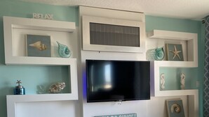 Una sala de estar con un televisor montado en un mueble blanco, estantes con objetos decorativos y un letrero que dice "LET'S DO SUNSET".
