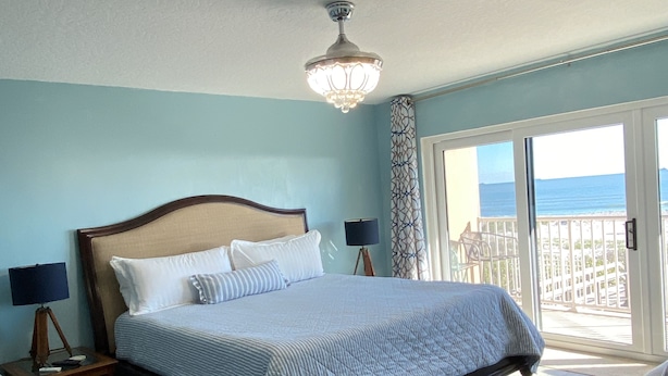 Un dormitorio con cama, mesita de noche, lámpara, silla y vista a la playa a través de la ventana.
