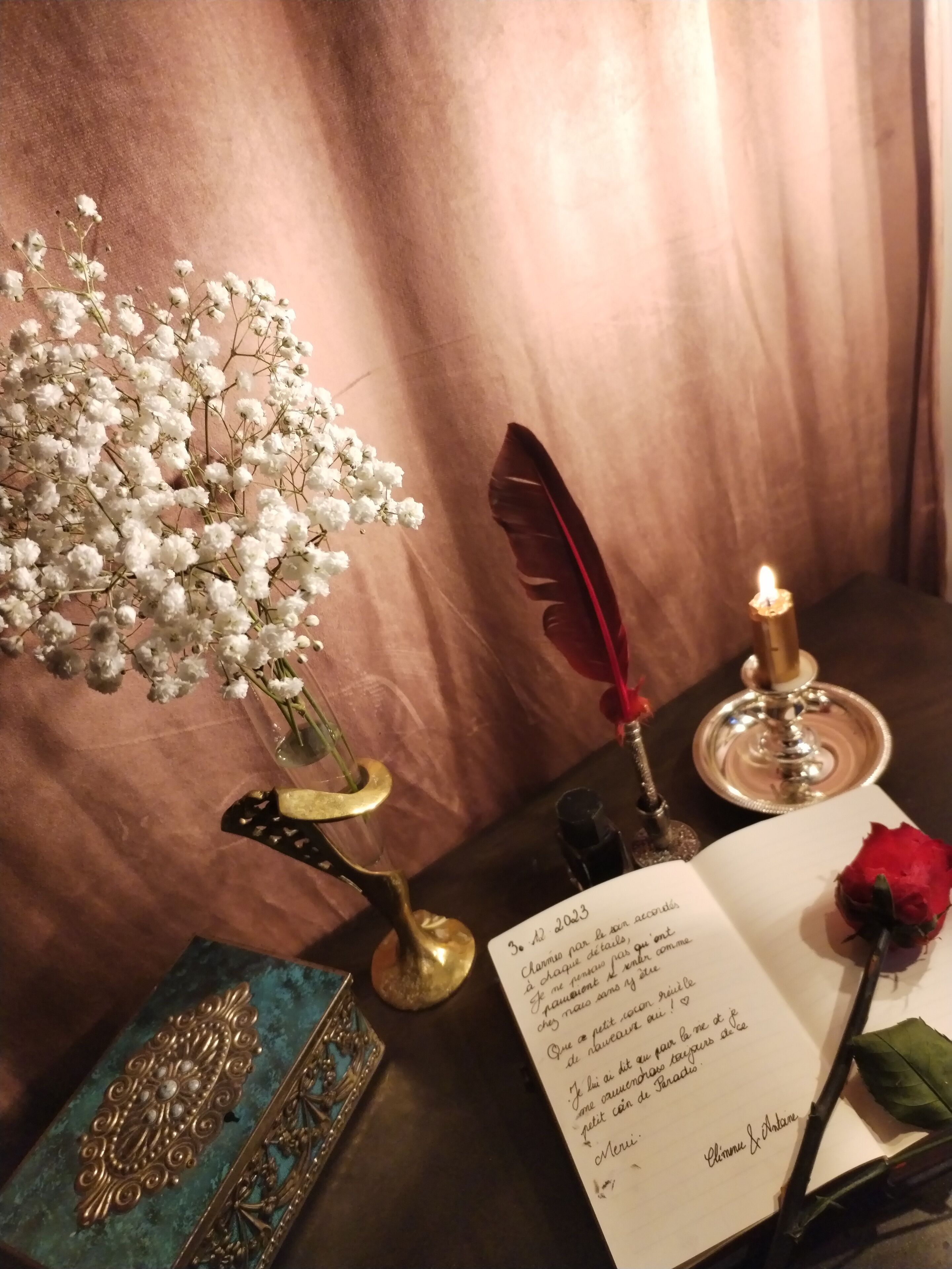 Une table ornée d’un vase de fleurs blanches, d’une plume d’oie, d’une bougie, d’un livre et d’une rose rouge.