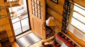 Interior acolhedor de uma cabana com piso de madeira, cama com roupa de cama estampada, porta que dá para o exterior e uma pequena mesa com abajur.