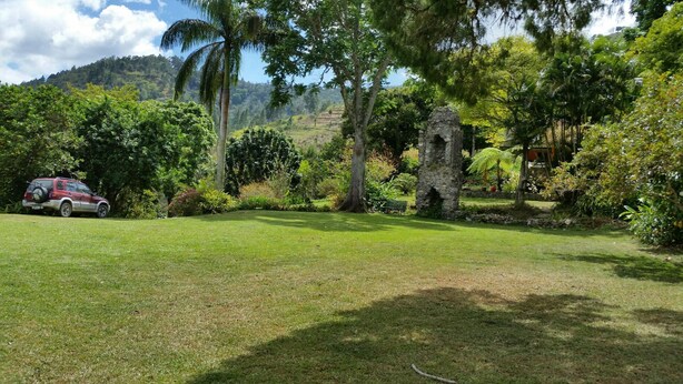 A grassy area with a stone structure, trees, and a parked car. - Blue Mountains Jamaican Vacation (St Peters)