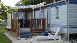 En campingvogn med veranda, en hvid liggestol og et lille haveområde med plantekasser.