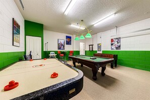 A pool table, a foosball table, and a dartboard in a room with green and white walls, a ceiling fan, and sports-themed decorations.