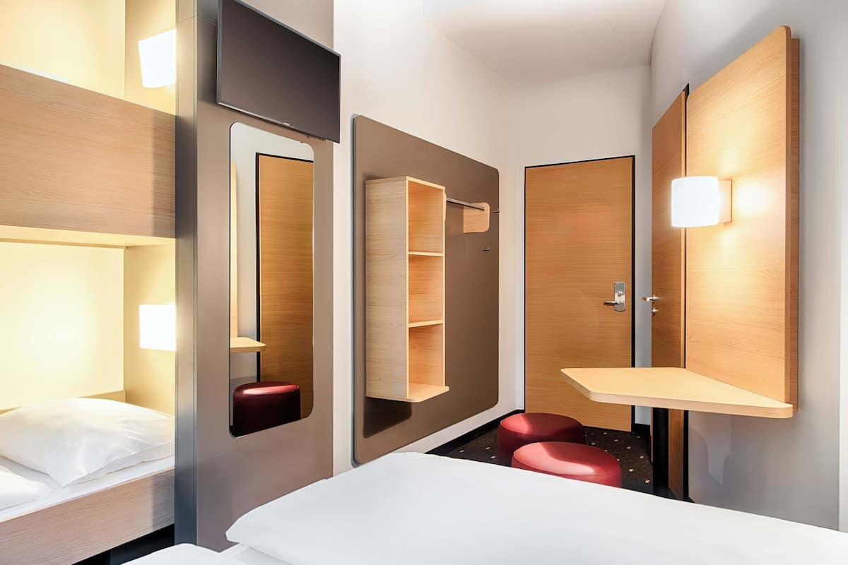 Quarto de hotel moderno com uma cama, um espelho grande, um guarda-roupa de madeira e um banquinho vermelho pequeno.