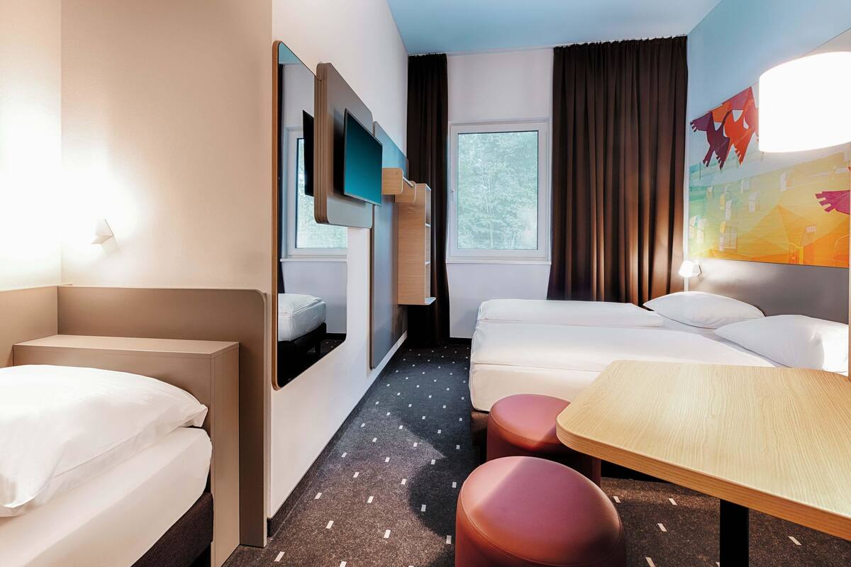 Quarto de hotel com cama, escrivaninha, cadeira, televisão e janela.