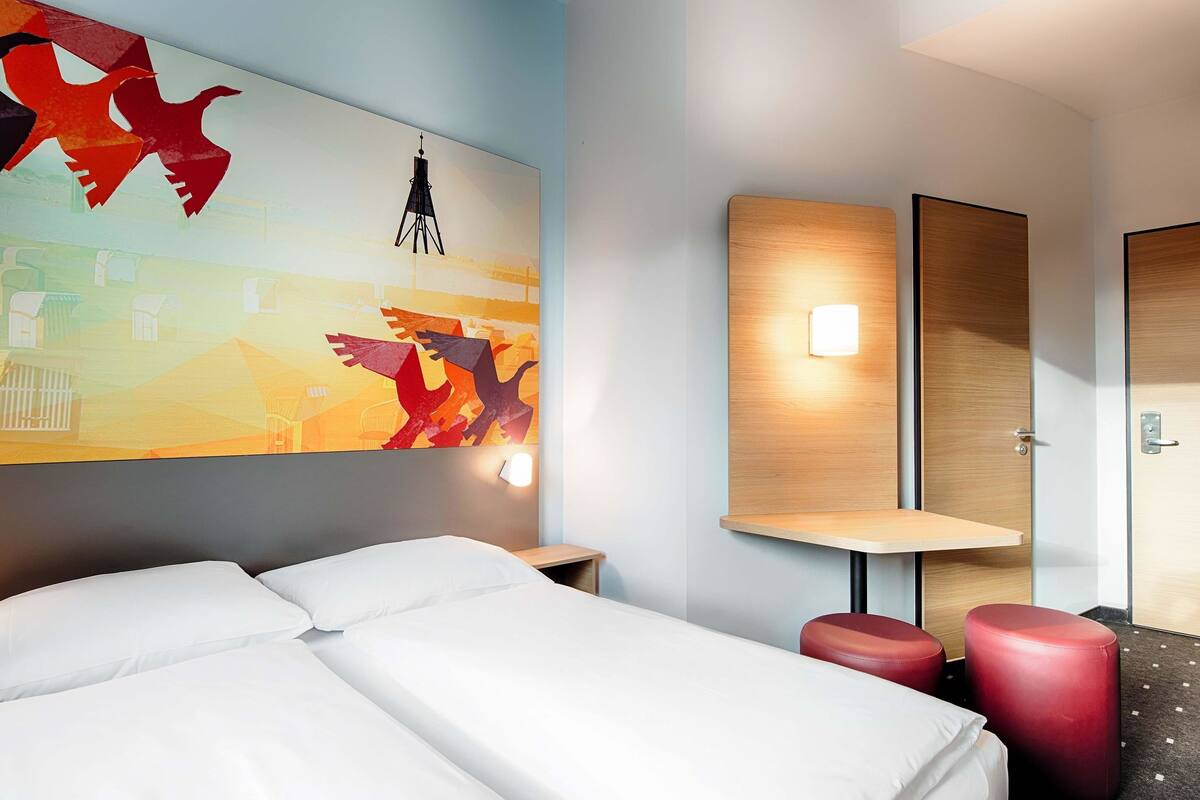 Quarto de hotel com cama, um banquinho vermelho, luz acoplada à parede e uma grande pintura abstrata.