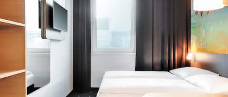 Quarto de hotel com cama, televisão fixada na parede, janela com cortinas e um quadro na parede.