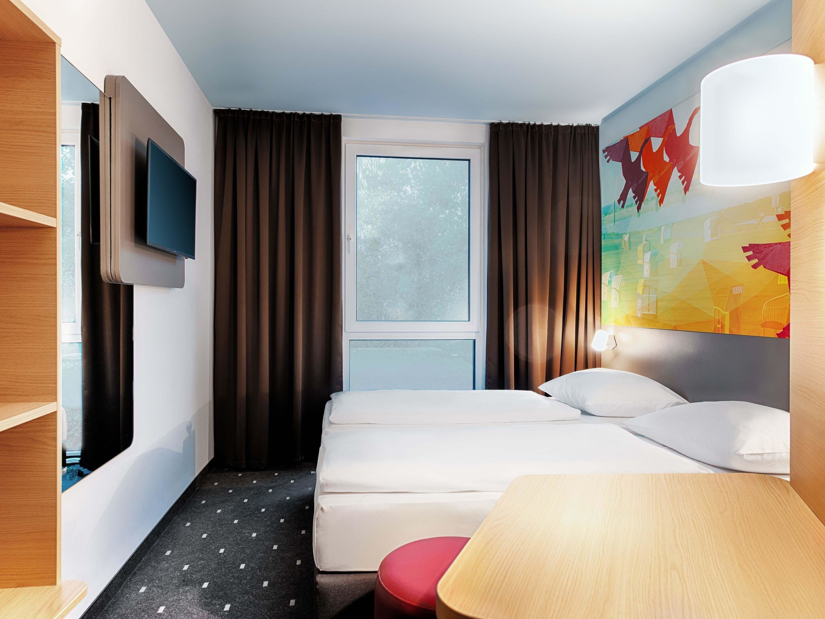 Una habitación de hotel con cama, televisor montado en la pared, ventana con cortinas, un cuadro y una lámpara.
