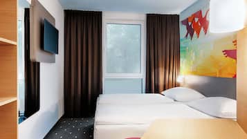 Una habitación de hotel con cama, televisor montado en la pared, ventana con cortinas, un cuadro y una lámpara.