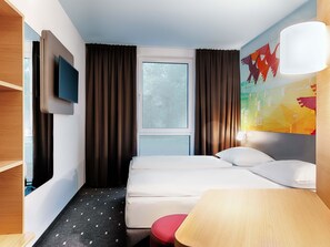 Una habitación de hotel con cama, televisor montado en la pared, ventana con cortinas, un cuadro y una lámpara.