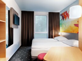 Une chambre d’hôtel avec un lit, un téléviseur fixé au mur, une fenêtre avec des rideaux, un tableau au mur et une lampe.