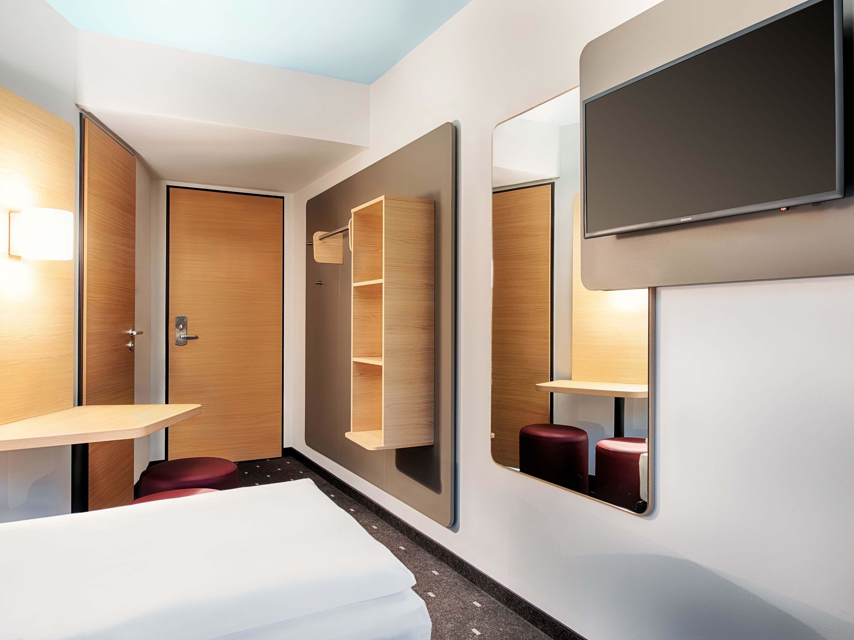 Een moderne hotelkamer met een bed, een aan de muur bevestigde televisie, een houten garderobe en een klein tafeltje.
