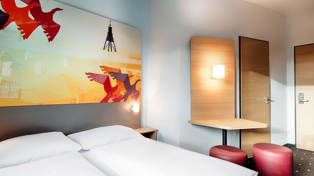 Habitación de hotel con cama, un reposapiés rojo, una lámpara de pared y un cuadro abstracto grande.