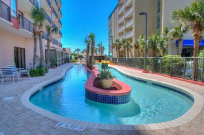 A swimming pool with a tiled hot tub, surrounded by palm trees and a building with balconies.