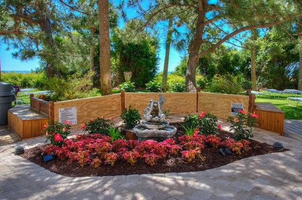 Un jardín ajardinado con una fuente central, rodeado de bancos de madera y flores coloridas.