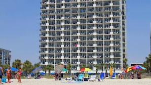 Ein mehrstöckiges Hotel mit Balkonen, direkt an einem Strand mit Liegestühlen und Sonnenschirmen.