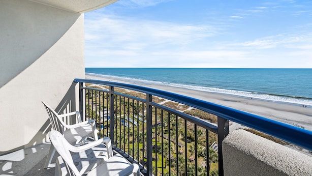 Ein Balkon mit weißen Plastikstühlen, Blick auf den Strand und einem klaren, blauen Himmel.
