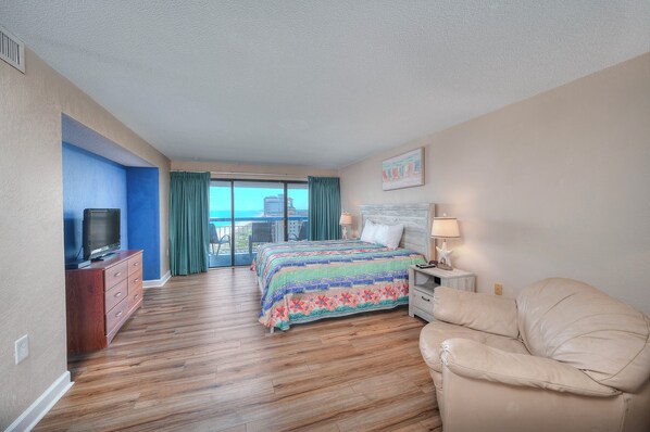 A hotel room with a large bed, a TV, a leather armchair, and a balcony with a view.