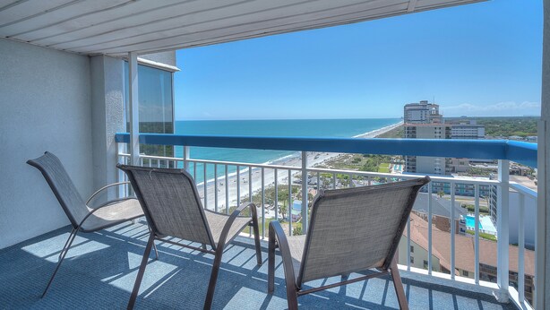 A balcony with two chairs, a view of the beach, and a building in the background.