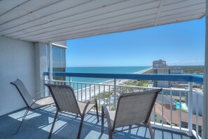 A balcony with two chairs, a view of the beach, and a building in the background.