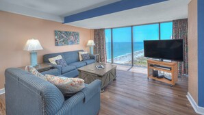 A living room with a large flat-screen TV, a sofa, a coffee table, and a view of the ocean.