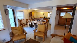 Amplia sala de estar con un sofá, sillas de madera y una mesita de centro. Se ve una cocina al fondo con una zona de comedor.