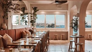 A restaurant with a long dining table, cushioned seating, and large windows offering a view of the city. - ★ Sunshine Loft With Balcony by ★ Sydney Dreams ★ (Bondi Beach)