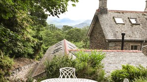 A patio with a round table and chairs, surrounded by greenery, with a stone building in the background. - Norton (Keswick)