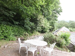 Une terrasse extérieure avec une table ronde et des chaises blanches, entourée d’une végétation luxuriante et d’un mur de pierre.