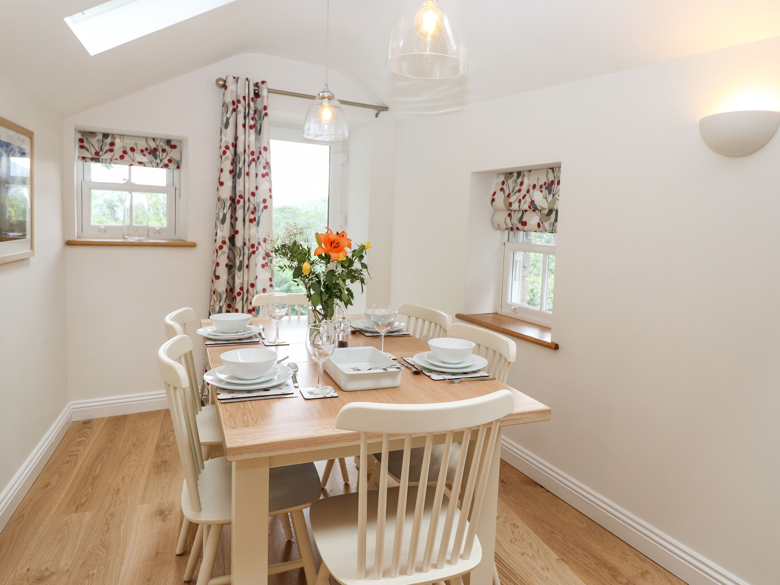 A dining area with a wooden table set for four, white chairs, and a vase of orange flowers.
