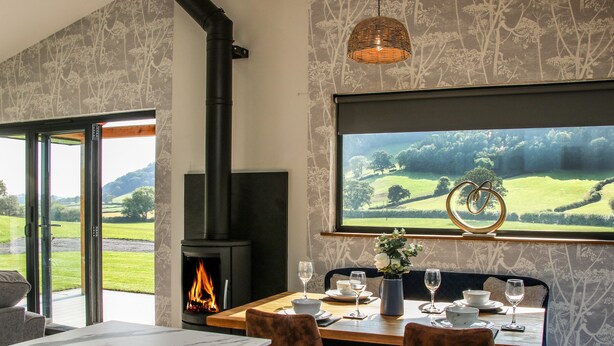 A modern kitchen with a built-in fireplace, a dining area with two chairs, and a large window with a scenic view. - Meadow View (Meifod)