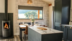 A modern kitchen with a wood-burning stove, a dining area with a view of the outdoors, and dark cabinetry. - Meadow View (Meifod)