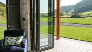 A modern room with a sliding glass door leading to a deck with a scenic view of a green landscape. - Meadow View (Meifod)