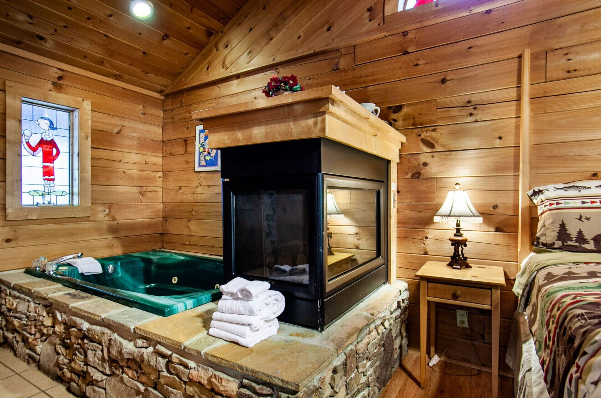 Interior de una cabaña de madera con chimenea de piedra, un jacuzzi verde y una cama con manta de temática forestal.