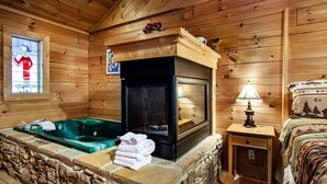 A wooden cabin interior with a stone fireplace, a green hot tub, and a bed with a forest-themed blanket.