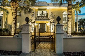 Una entrada cerrada con un portón de hierro forjado, un barandal de piedra y un camino flanqueado por palmeras que conduce a un edificio con acceso de cerámica.