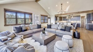 A spacious living room with a large sectional sofa, a coffee table, and ottomans. There is a kitchen area visible in the background. - Cocoa Cabin: New Baldy Mtn Home, Hot Tub & Steam Shower, EV Charger! (Breckenridge)