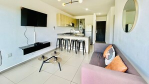 Un salon moderne doté d’un téléviseur à écran plat, d’un canapé, d’une table basse et d’un coin cuisine avec des tabourets de bar.