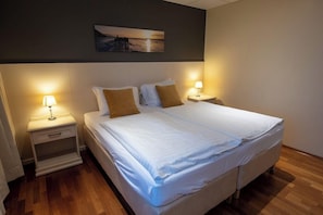 A neatly made bed with white linens and two pillows, flanked by bedside tables with lamps, in a room with wooden flooring and a painting on the wall. - Sel-hotel Myvatn (Myvatn)