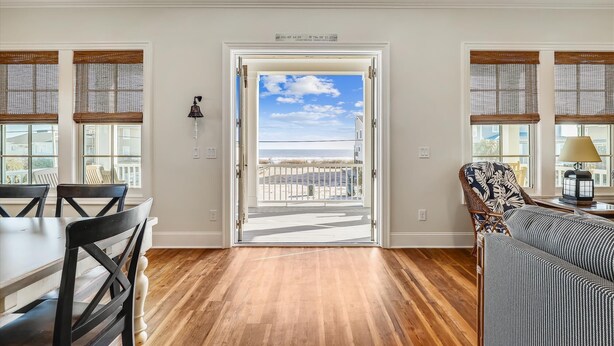 A room with a wooden floor, a dining table with chairs, a patterned armchair, and a view of the beach through a door.