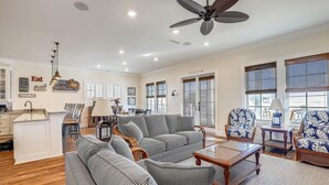 A spacious living room with a large sofa, a coffee table, and a ceiling fan. There is a dining area with a table and chairs, and a kitchen area with cabinets and a sink.