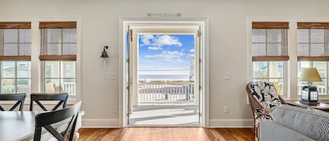 Una habitación con piso de madera, una mesa de comedor con sillas, un sillón estampado y vista a la playa a través de una puerta.