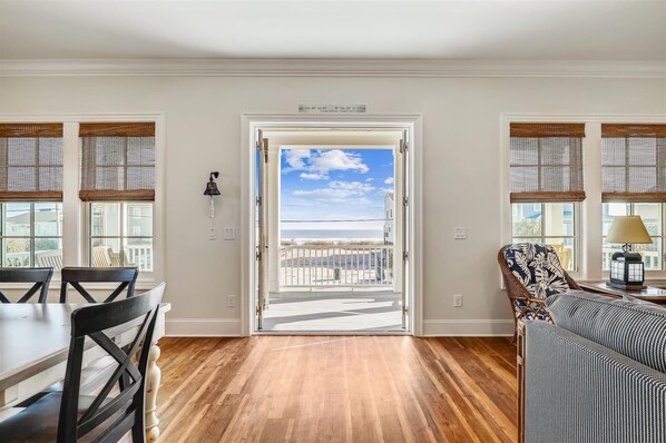 A room with a wooden floor, a dining table with chairs, a patterned armchair, and a view of the beach through a door.