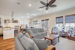 A spacious living room with a large sofa, a coffee table, and a ceiling fan. There is a dining area with a table and chairs, and a kitchen area with cabinets and a sink.