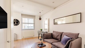 A living room with a brown sofa, a round wooden coffee table, a black clock, and a large mirror on the wall. - Soho by Excel Apartments (London)