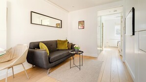 A modern living room with a grey sofa, a small side table, and a chair. There is a mirror on the wall and a door leading to another room. - Soho by Excel Apartments (London)
