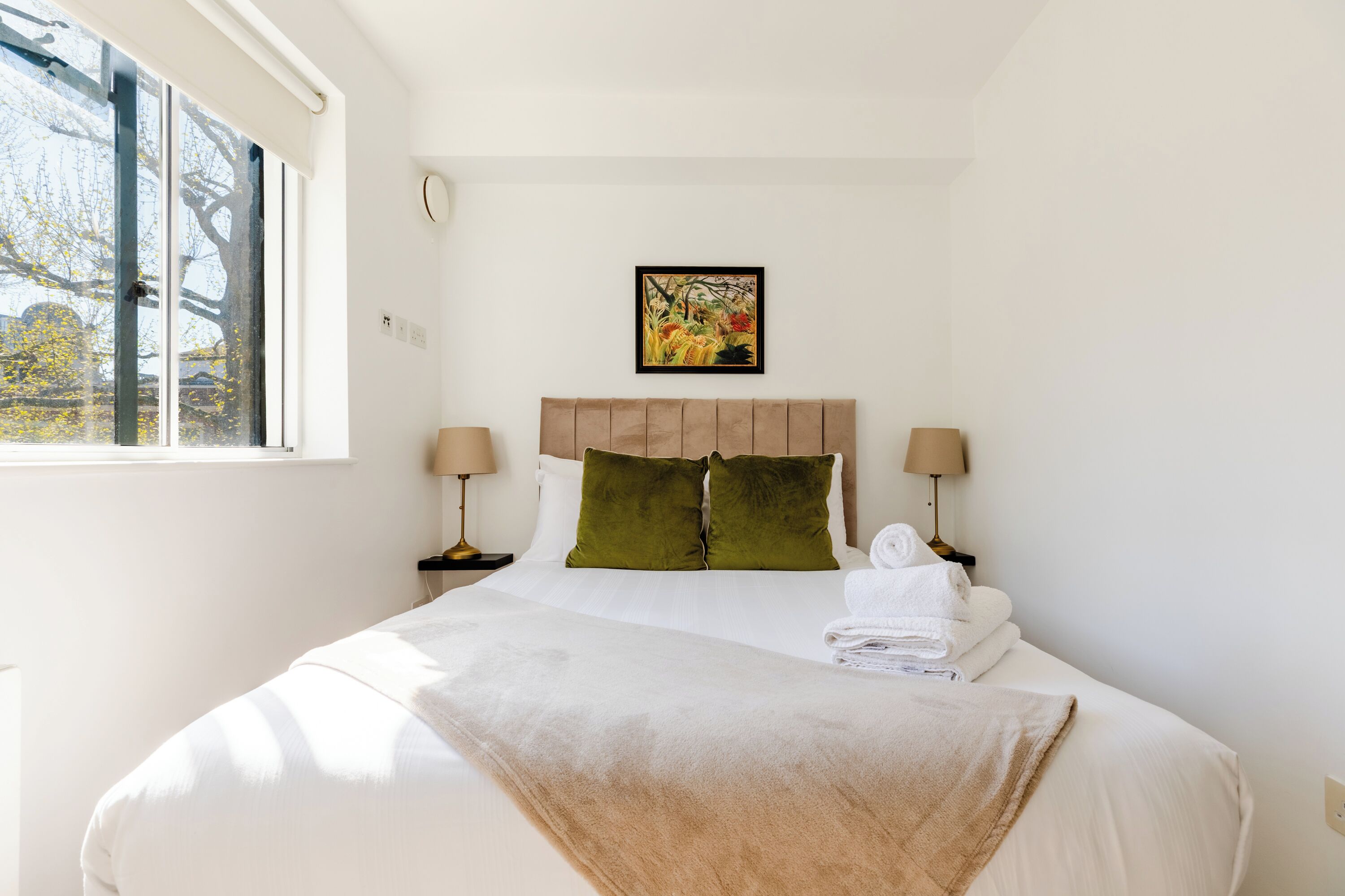 A neatly made bed with a beige blanket, green pillows, and white linens. There are two bedside lamps and a framed floral artwork above the bed.